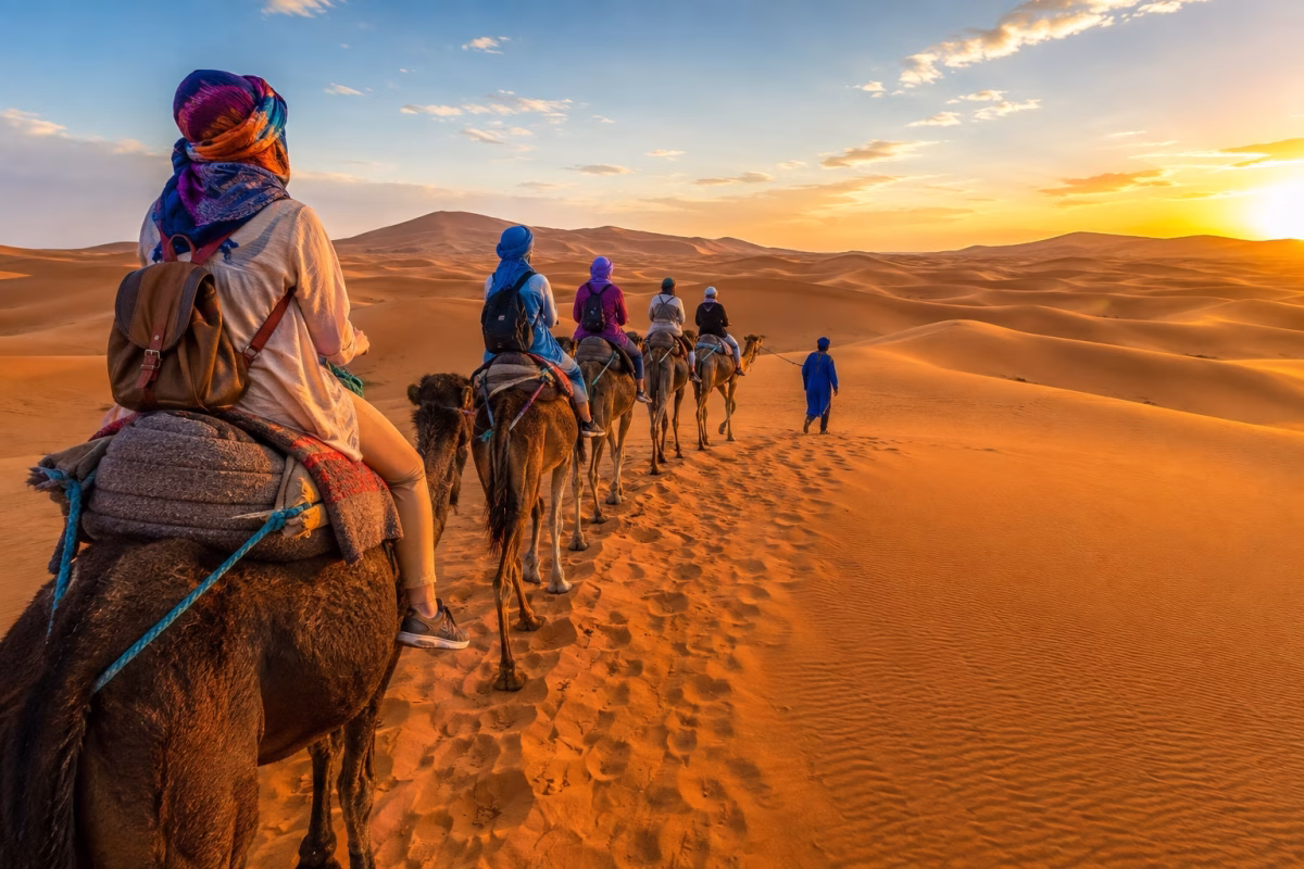 Travelers riding camels across the golden Erg Chebbi dunes at sunset during a Sahara desert tour from Tangier